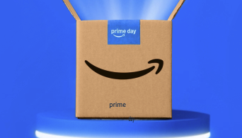 The image shows a cardboard box with the Amazon logo and the words "prime day" on a blue label. The box is open and placed on a blue platform with a glowing edge, set against a blue background with light beams emanating from the box.